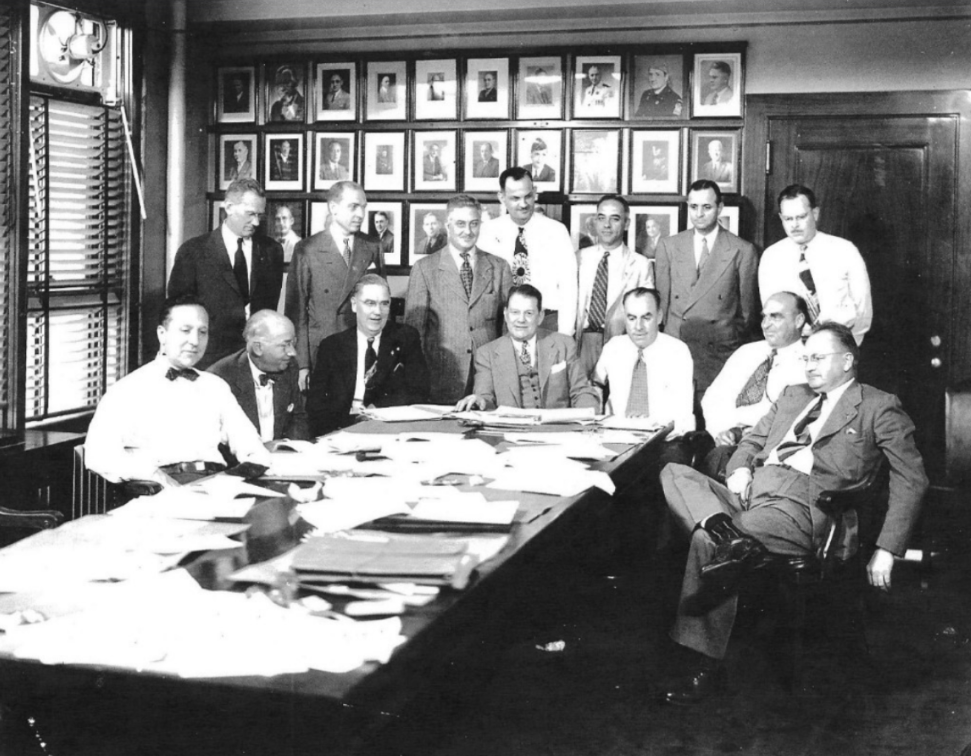 Historic black-and-white photograph of a group of fourteen people gathered around a long conference table in an office or boardroom. Several individuals are seated at the table while others stand behind them. The tabletop is covered with papers, folders, and documents, suggesting a business meeting or planning session. The room features wood-paneled doors, tall windows with blinds, and a large display of framed portraits arranged in rows on the back wall. The participants are dressed in formal business attire typical of the early to mid-twentieth century.