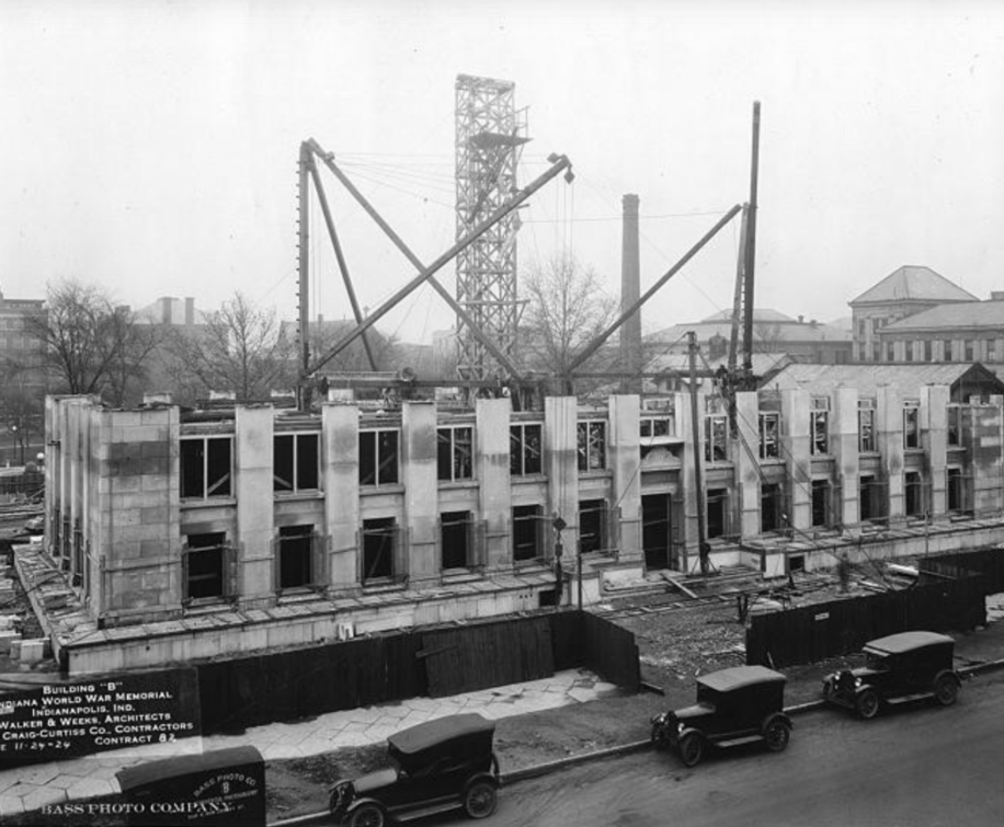 Historic black-and-white photograph showing construction of Building “B” of the Indiana World War Memorial in Indianapolis. A partially completed stone building occupies the foreground, with open window bays and unfinished upper walls exposing the building's structural framework.
