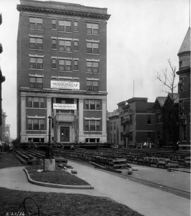 Historic black-and-white photograph of a six-story brick commercial building standing along a city street. The building features a symmetrical façade with rows of rectangular windows, decorative stone lintels, and a projecting cornice along the roofline. A sign mounted above the main entrance advertises office space, while another sign identifies a business located on the lower floors. In the foreground, steel beams and construction materials are laid out in parallel rows across a cleared section of roadway, indicating an active construction project. Residential buildings line the street to the right, including a house with a turret and several porches. A small group of people stands on the sidewalk in the background observing the work. The photograph is dated August 21, 1926, in the lower left corner.
