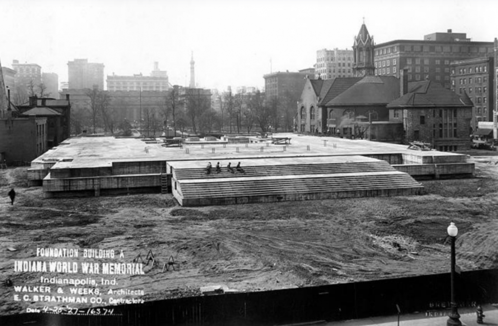 Historic black-and-white photograph showing the foundation construction of the Indiana World War Memorial in Indianapolis. A large rectangular concrete foundation fills the center of the image, with broad stepped sections visible along the near side. Workers and construction materials are scattered across the site, which is surrounded by cleared earth and temporary fencing.