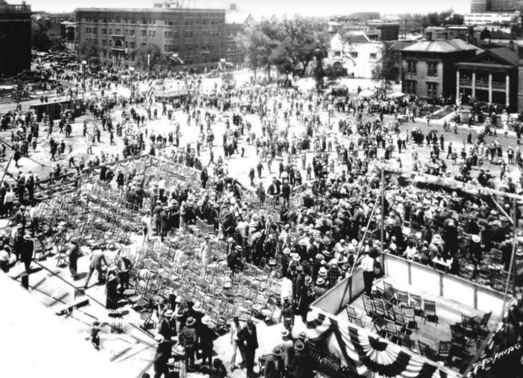 Historic black-and-white photograph showing a large outdoor gathering at what appears to be a ceremonial or civic event. Hundreds of people are assembled around a temporary stage and seating area decorated with patriotic bunting.