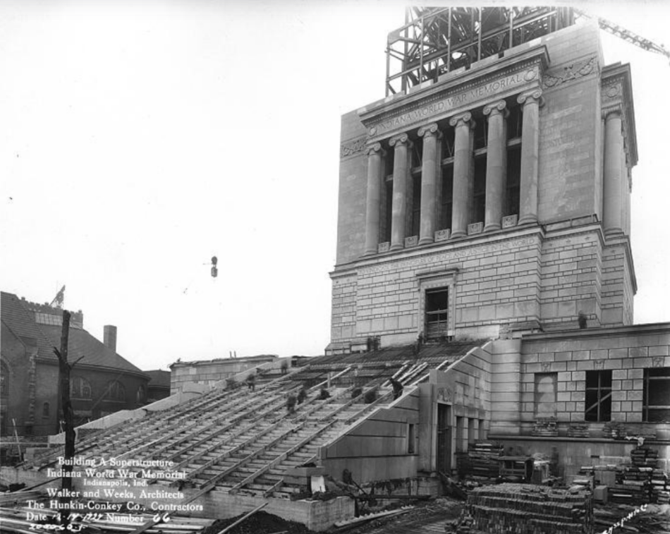 Historic black-and-white photograph of the Indiana World War Memorial during construction. The monumental neoclassical building dominates the scene, with a tall columned upper structure rising above a broad stone base. The words “Indiana World War Memorial” are carved into the entablature above the columns. Temporary wooden forms and scaffolding cover the wide stairway leading to the main entrance, indicating that construction was still in progress.