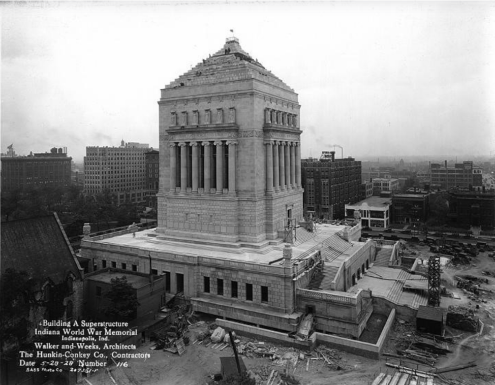 Historic black-and-white photograph of the nearly completed Indiana World War Memorial in Indianapolis. The monumental neoclassical structure rises above its surrounding base, featuring tall fluted columns on all sides and a stepped pyramidal roof topped with a small flag. Broad stairways and terraces surround the building, while construction materials, equipment, and unfinished landscaping remain visible around the site.