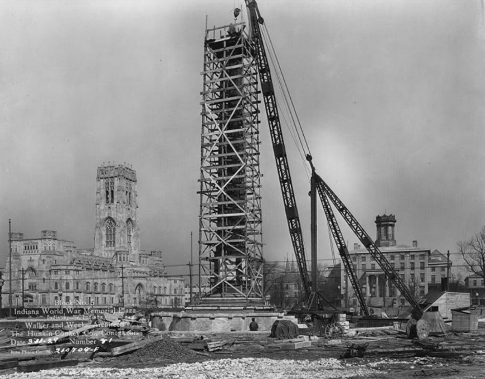 Historic black-and-white photograph showing construction of the Indiana World War Memorial Plaza obelisk in Indianapolis. A tall steel framework marking the future monument rises from a stone base at the center of an active construction site. Large cranes and lifting equipment surround the structure, with construction materials scattered across the foreground.