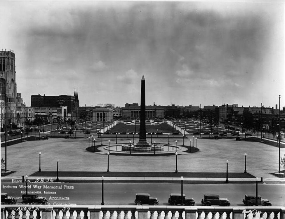 Historic black-and-white photograph of the completed Indiana World War Memorial Plaza in Indianapolis. A tall obelisk monument stands at the center of a circular plaza surrounded by a low fountain or basin and evenly spaced lampposts.