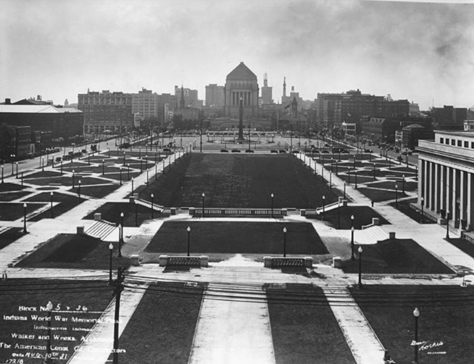 Historic black-and-white photograph of the completed Indiana World War Memorial Plaza in Indianapolis. The image looks south along a formal, symmetrical landscape composed of broad lawns, paved walkways, terraces, stairways, and evenly spaced lampposts.