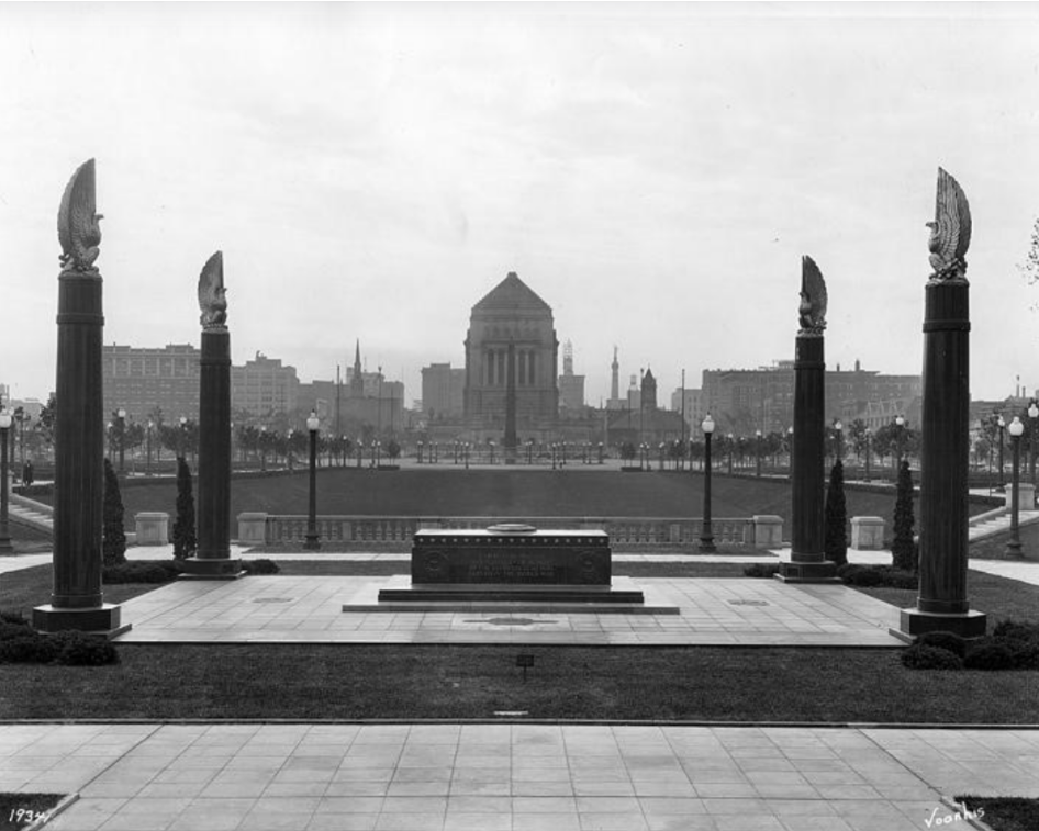 Historic black-and-white photograph of a memorial terrace overlooking the Indiana World War Memorial Plaza. Four tall columns topped with sculptural eagles frame a central stone monument or cenotaph in the foreground. Beyond the terrace, the landscaped plaza stretches toward the Indiana World War Memorial building in the distance.