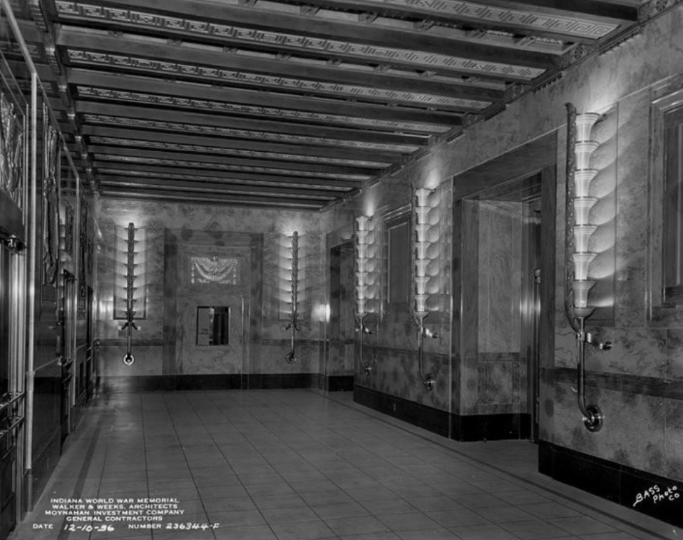 Historic black-and-white photograph of an interior corridor in the Indiana World War Memorial. The hallway features marble walls, a decorative coffered ceiling, and a series of illuminated Art Deco-style wall sconces mounted along both sides. Large doorways and decorative stone trim line the corridor, while a tiled floor extends toward a doorway at the far end of the hall.