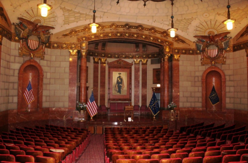 Interior view of an ornate auditorium in the Indiana World War Memorial. Rows of red upholstered theater seats face a raised stage framed by red marble columns, decorative gold detailing, and patriotic eagle motifs. An American flag and an Indiana state flag stand on either side of the stage, while a large portrait hangs at the center of the back wall. Pendant lights hang from the domed, elaborately decorated ceiling above the seating area.