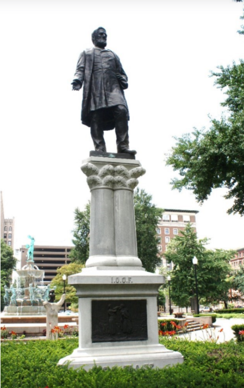 Color photograph of the Schuyler Colfax monument in a landscaped city park. A bronze standing figure is mounted atop a tall stone pedestal supported by clustered columns. The pedestal is inscribed “I.O.O.F.” and includes a bronze relief panel. Flower beds and trimmed shrubs surround the monument, while a decorative fountain, trees, and downtown buildings are visible in the background.