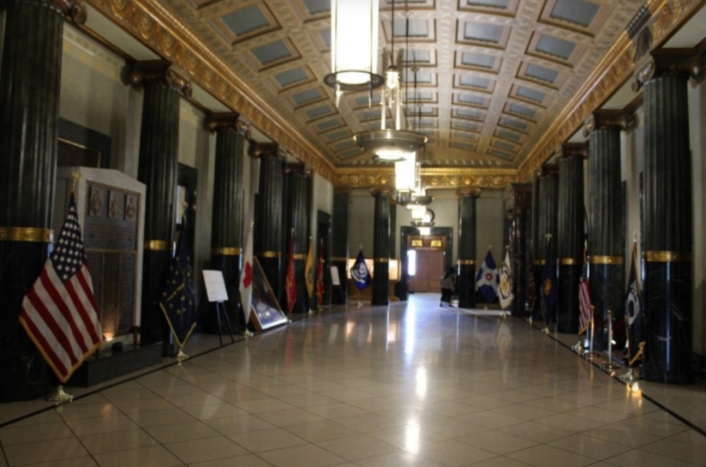 Interior view of the Indiana World War Memorial foyer featuring polished marble columns, a coffered ceiling with decorative gold detailing, and a wide tiled floor. American, military, and state flags line both sides of the hall, while memorial plaques and exhibits are displayed along the walls. Large pendant light fixtures hang from the ceiling, leading the eye toward doors at the far end of the space.