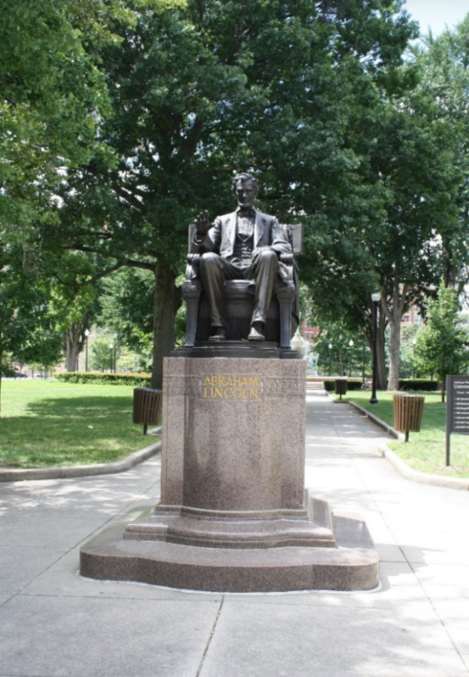 Color photograph of a seated Abraham Lincoln statue mounted on a polished granite pedestal engraved with the name “ABRAHAM LINCOLN” in gold lettering. The bronze figure sits in a chair facing forward along a paved park walkway. Mature trees, manicured lawns, benches, and informational signs surround the monument, creating a shaded park setting.