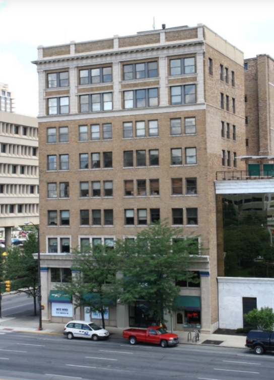 Modern photograph of the Medical Arts Building, a mid-rise brick commercial building with large rectangular windows and decorative stone trim. The seven-story structure fronts a city street lined with trees, where several parked vehicles are visible along the curb.