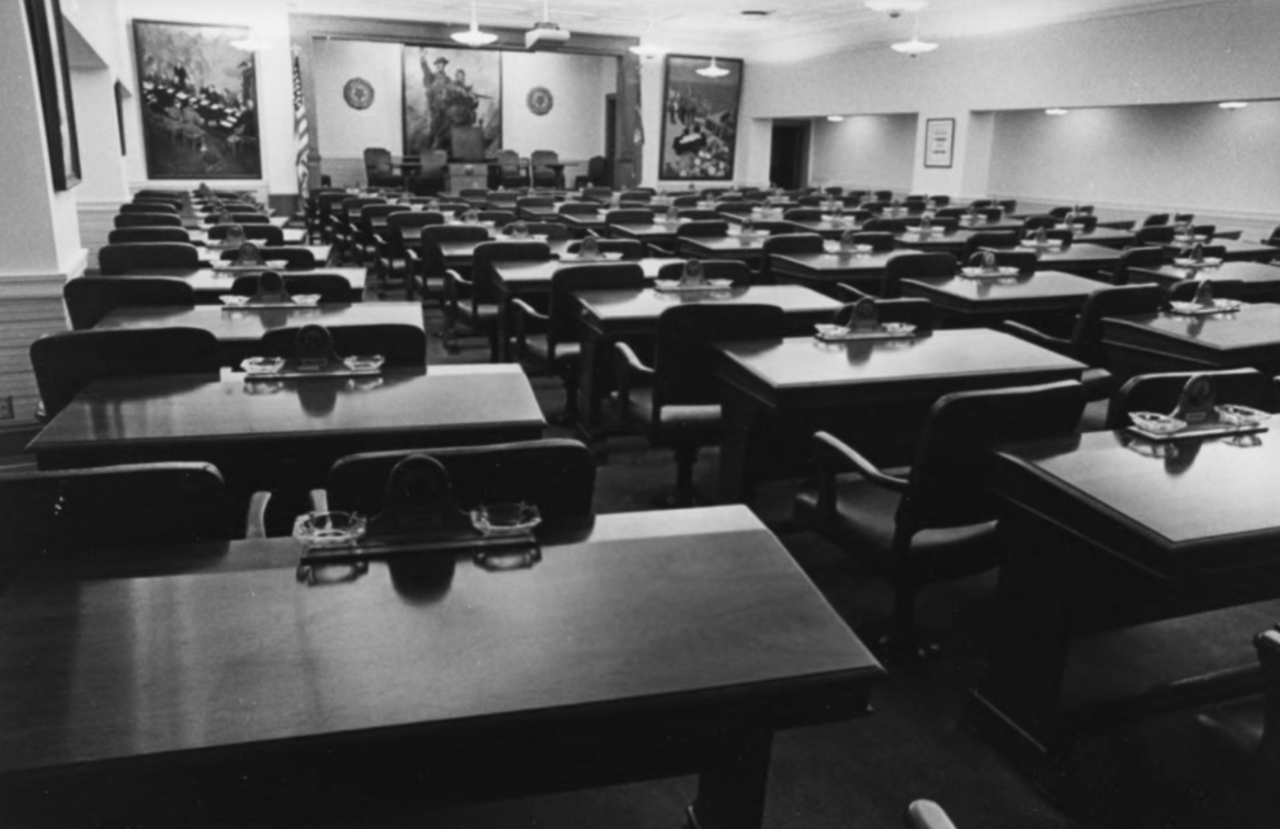 Interior view of a large meeting chamber with rows of rectangular desks and chairs arranged to face a raised dais at the front of the room. Flags, large framed murals, and official seals decorate the front wall, creating a formal governmental or civic setting.