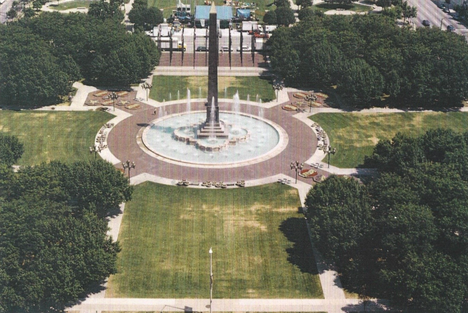 Aerial view of a formal park centered on a tall dark obelisk rising from a large circular fountain. Brick walkways radiate outward from the fountain in a symmetrical pattern, dividing broad lawns and rows of mature trees. Benches and flower beds line the paths, and city streets and buildings are visible beyond the tree-lined park.