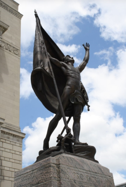 Color photograph of a bronze allegorical statue standing atop a granite pedestal. The figure holds a large flag that billows behind them and raises one arm upward. The statue is set against a partly cloudy blue sky, with the stone facade of a monumental building visible along the left edge of the image. The base of the monument includes the inscription “PRO PATRIA."