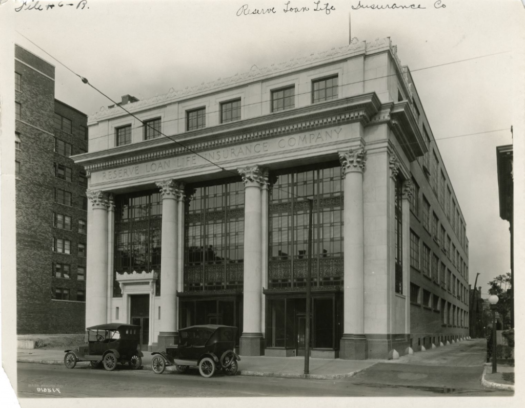 Historic black-and-white photograph of the Reserve Loan Life Insurance Company building, a neoclassical commercial structure with a monumental façade featuring four large Corinthian columns. The building occupies a street corner and displays the company name across the entablature above tall multi-story windows