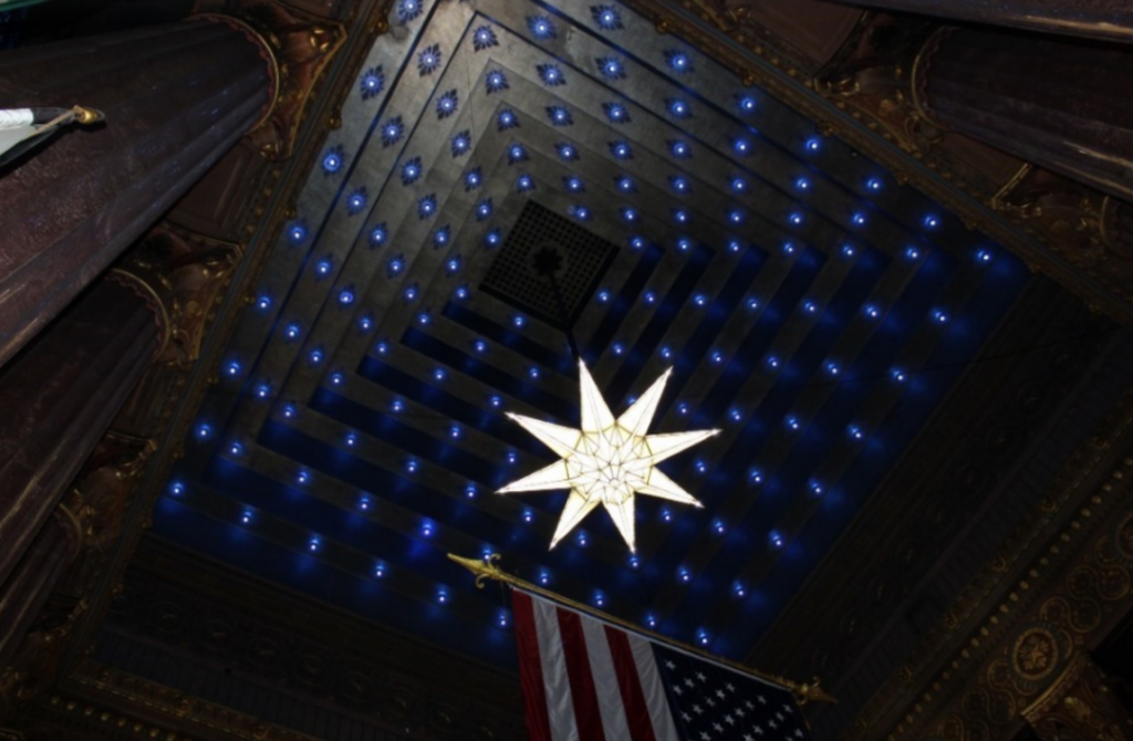 Upward view of the Indiana World War Memorial Shrine Room ceiling, featuring a large illuminated star-shaped chandelier suspended beneath a stepped ceiling filled with small blue lights resembling stars.
