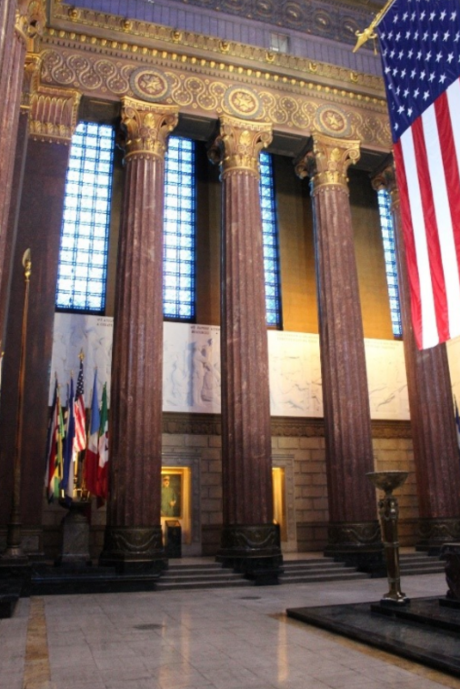 Interior view of the Indiana World War Memorial Shrine Room featuring tall marble columns with ornate capitals, blue stained-glass windows, marble relief panels, and a large American flag hanging from the ceiling. The grand ceremonial space is illuminated by natural light filtering through the towering windows.