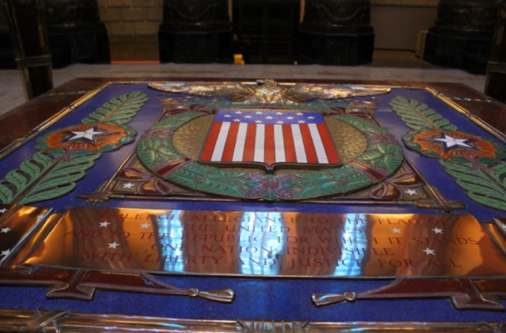 Close-up view of an ornate memorial plaque or floor panel featuring a large United States coat of arms at its center. The design includes a red, white, and blue shield beneath a gold eagle with outstretched wings, surrounded by decorative green laurel branches, stars, and military-themed motifs.