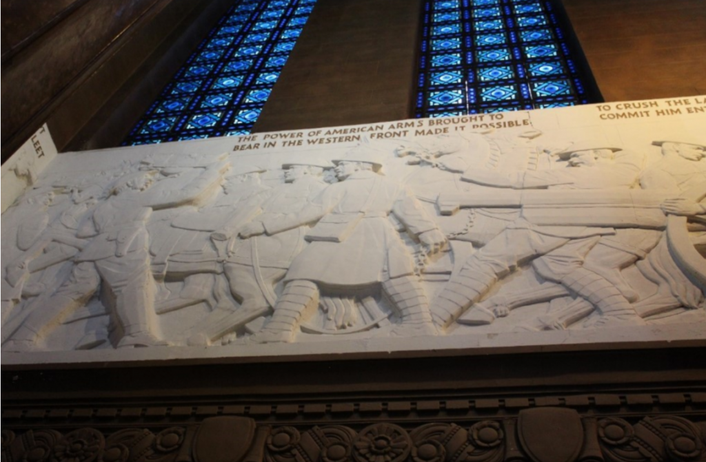 Interior view of a monumental memorial hall featuring a large carved stone relief mounted high on a wall beneath tall stained-glass windows. The white bas-relief depicts uniformed soldiers advancing with military equipment, weapons, and artillery in a dynamic wartime scene. Portions of an inscription are visible above the relief, including the phrase, “THE POWER OF AMERICAN ARMS BROUGHT TO BEAR IN THE WESTERN FRONT MADE IT POSSIBLE.”