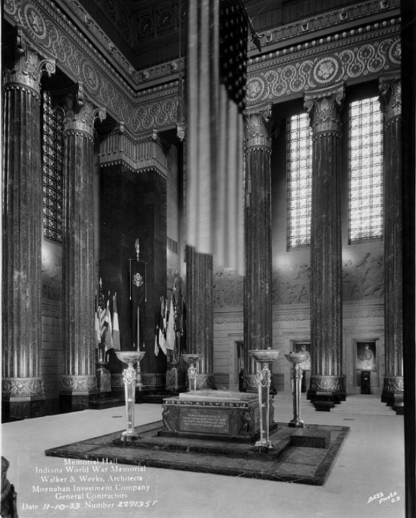 Historic black-and-white photograph of the Indiana World War Memorial Shrine Room. A large American flag hangs above a central stone altar or memorial block surrounded by decorative lampstands. Tall marble columns, stained-glass windows, and carved wall reliefs line the monumental interior, with military and national flags displayed along the far wall.