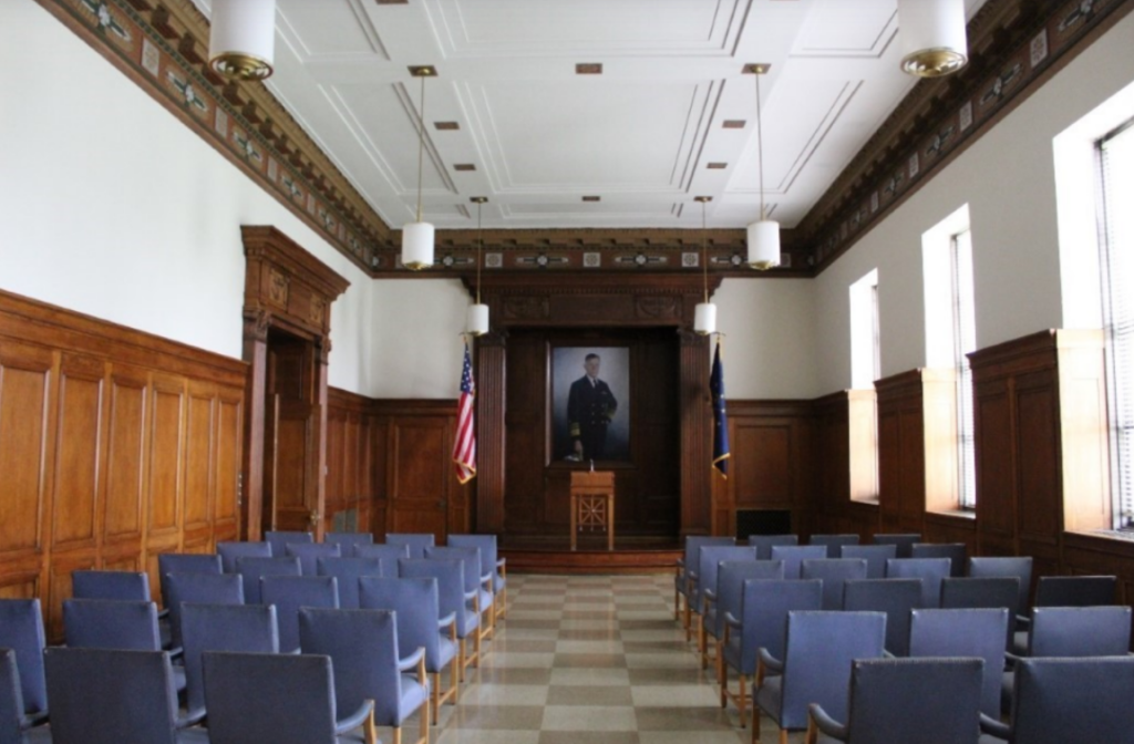Interior view of a memorial meeting room with wood-paneled walls, a decorative coffered ceiling, and rows of blue upholstered chairs facing a small stage. A large portrait hangs at the front of the room between an American flag and an Indiana state flag. Tall windows along one wall provide natural light, while pendant light fixtures hang from the ceiling above the seating area.