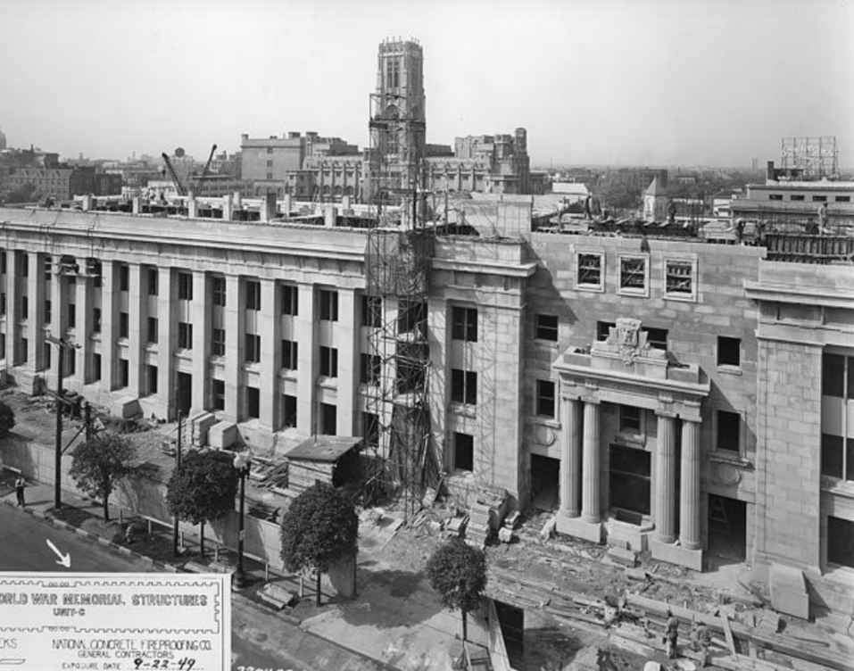 Exterior view of the Indiana World War Memorial during construction, featuring a long neoclassical façade with tall columns, unfinished stonework, scaffolding, and work materials along the street-facing side of the building.