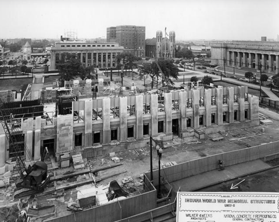 Elevated view of the American Legion under construction, showing a partially completed stone building with exposed structural supports, scaffolding, and construction materials scattered across the site. Streets, trees, parked cars, and neighboring downtown buildings surround the project, while a construction sign identifying the memorial is visible in the foreground.