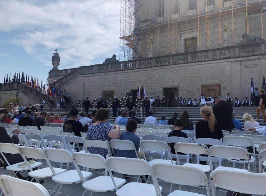 Color photograph of a public ceremony taking place on the steps of the Indiana World War Memorial. Rows of white folding chairs fill the foreground, with attendees seated and facing a group assembled on the memorial's broad staircase. Flags line the stair railings, floral wreaths are displayed near the speakers, and scaffolding is visible on the upper portions of the building, indicating ongoing restoration work. The event is held outdoors beneath a partly cloudy sky.