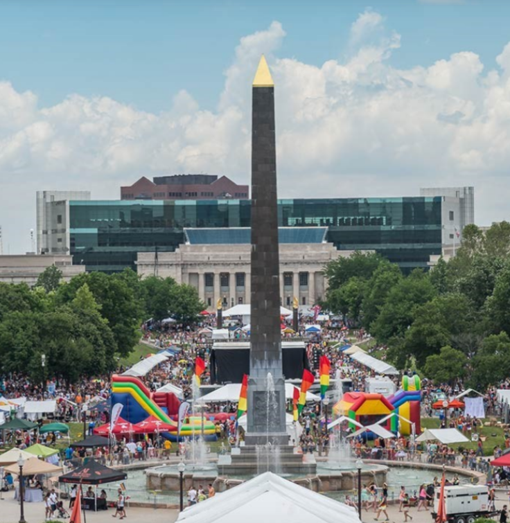 Aerial view of Veterans Memorial Plaza filled with people attending a large outdoor festival. The central black granite obelisk rises above a circular fountain surrounded by crowds, tents, and colorful inflatable attractions. Tree-lined pathways lead toward a large neoclassical building in the background, while modern office buildings frame the scene. The plaza is densely populated with visitors gathered around event spaces under a bright sky with scattered clouds.