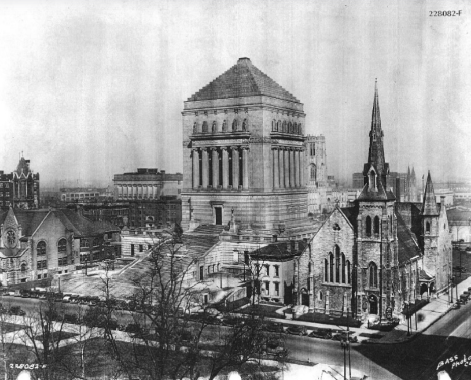 Black-and-white photograph of the Indiana World War Memorial rising above surrounding downtown Indianapolis buildings. The monumental stone memorial, topped with a pyramidal roof and lined with tall columns, dominates the center of the image. In the foreground, several church buildings with steep spires stand along nearby streets, while additional commercial buildings and city blocks extend into the background.