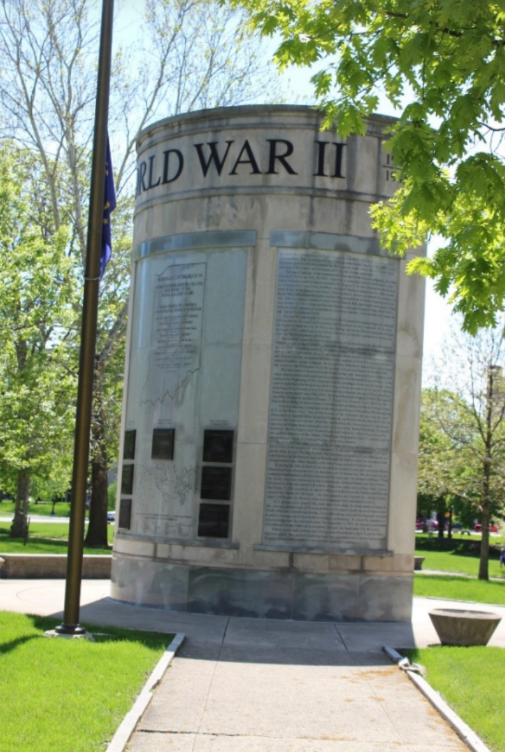 Color photograph of the Indiana World War II Memorial in a park setting. A tall cylindrical stone monument engraved with the words “WORLD WAR II” near the top stands at the end of a paved walkway. Large engraved panels listing names and commemorative text cover the surface of the memorial. A flagpole stands to the left, and green lawns, leafy trees, and pedestrian paths surround the monument under a clear blue sky.