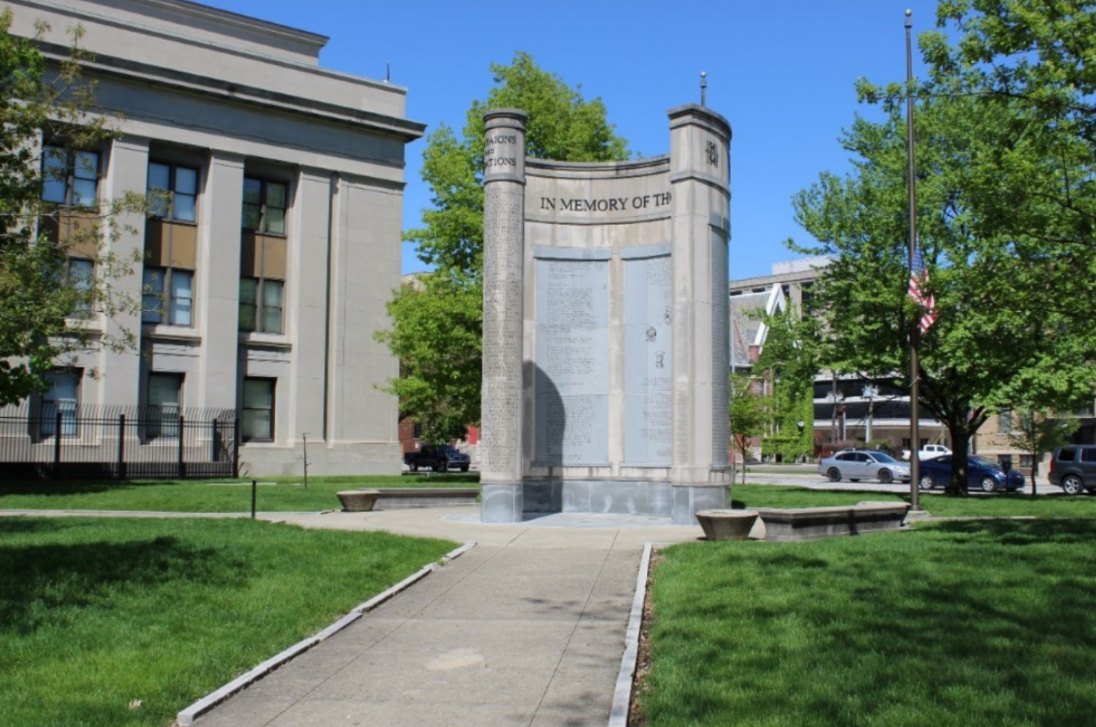 Color photograph of the Indiana World War II Memorial standing in a landscaped urban park. A curved stone monument with two cylindrical columns and engraved commemorative panels rises at the end of a paved walkway bordered by green lawns. The inscription “IN MEMORY OF THE” is visible across the upper portion of the memorial. A large civic building stands to the left, while trees, parked cars, and an American flag on a flagpole frame the scene under a bright blue sky.